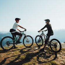 Two mountain bikers stand on a hillside looking at the landscape.
