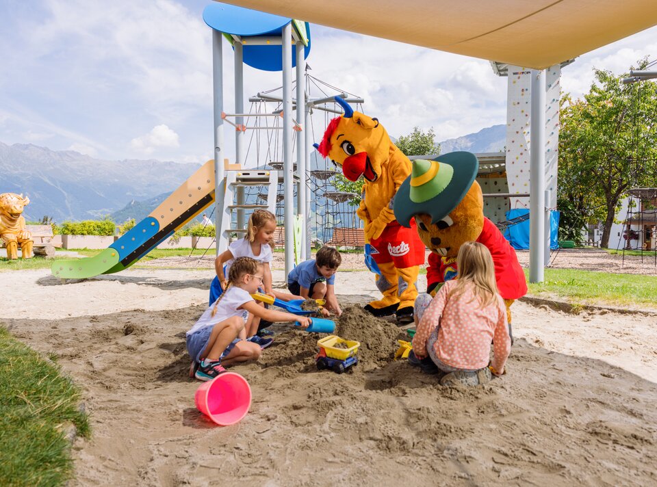 Kinder spielen im Sandkasten mit Maskottchen in einem Spielplatz.