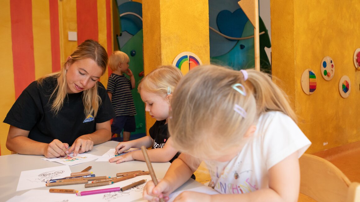 Een verzorger en twee kinderen zijn enthousiast aan het tekenen in een kleurrijke kamer.