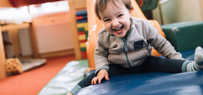 A happy toddler joyfully sits on a mat in a play area.