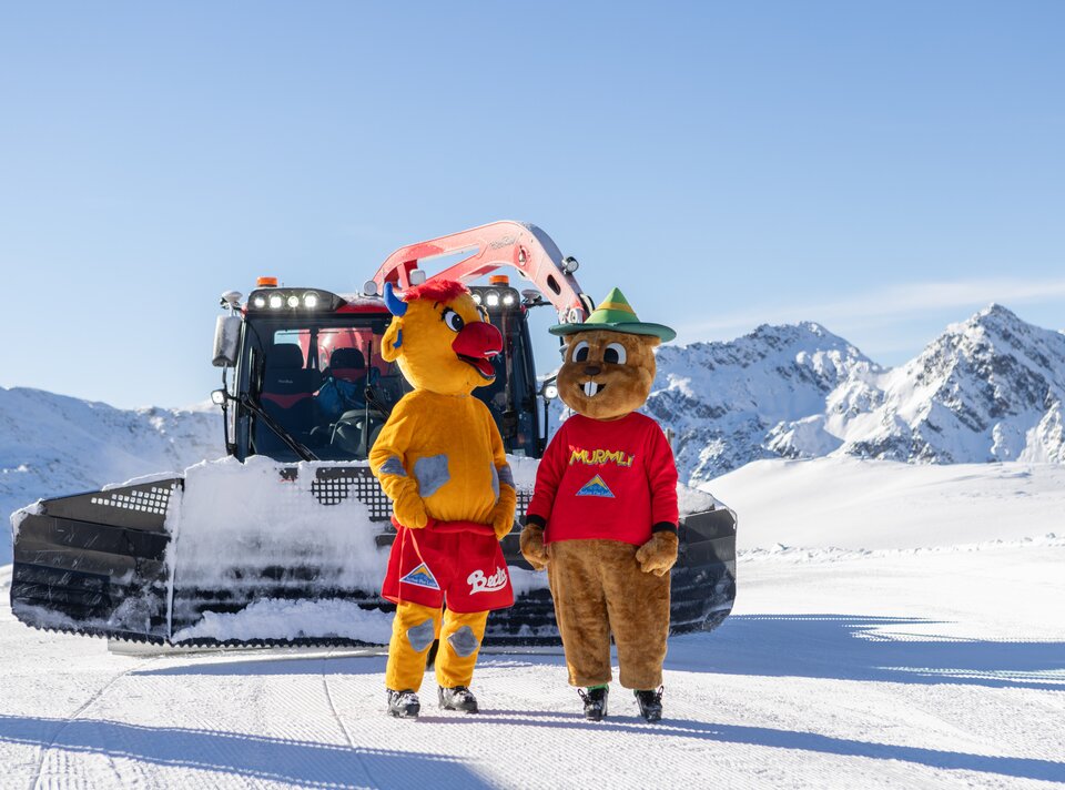 Zwei Maskottchen stehen im Schnee vor einem Pistenfahrzeug. Berge im Hintergrund.