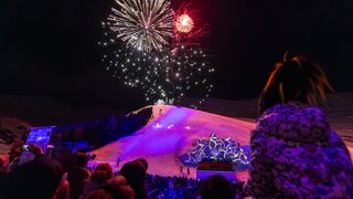 Feuerwerk erleuchtet den Nachthimmel über einer beleuchteten Skisprungveranstaltung.