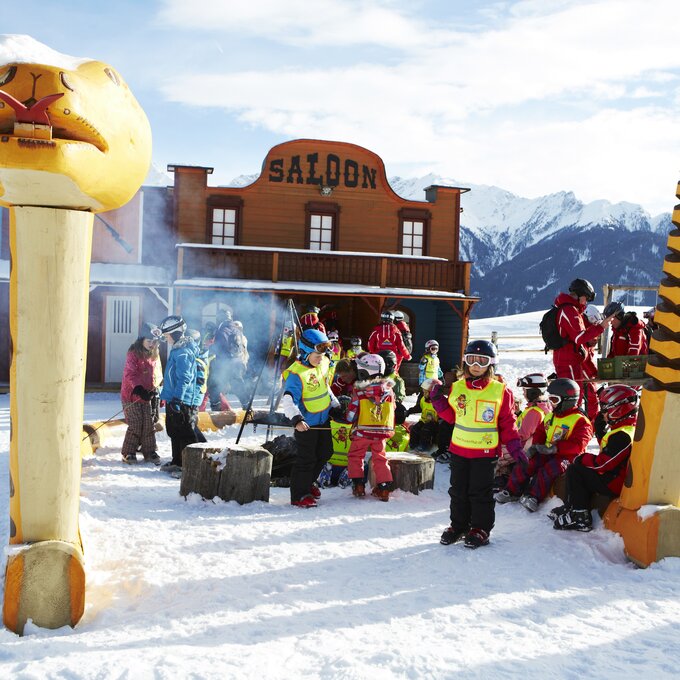 Gruppe von Kindern in Skikleidung versammelt vor einem Saloon im Schnee.