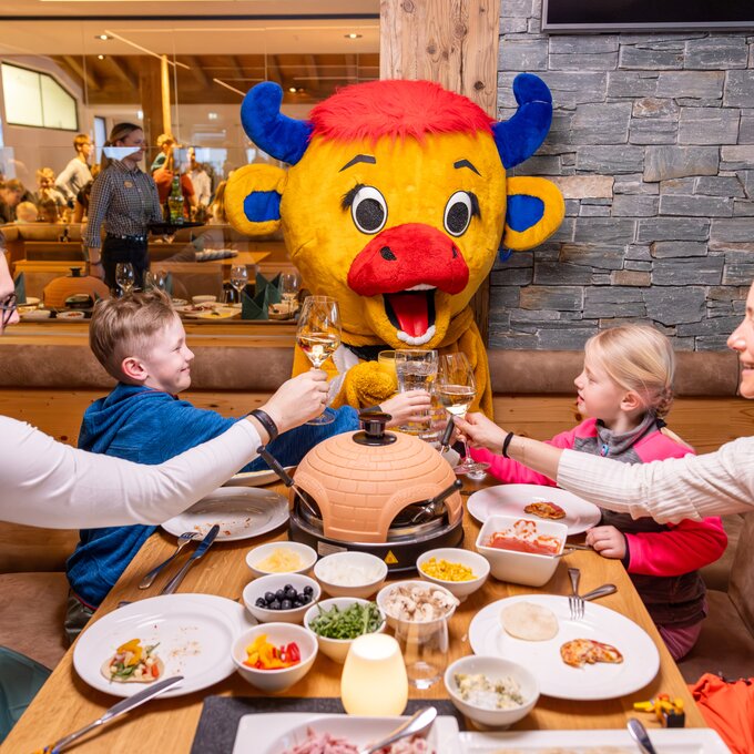 Familie stoßt mit Getränken an, während ein Maskottchen lächelt.