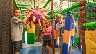 Four children play in a colorful indoor playground with large, soft toys.