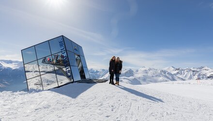 Two people in front of a reflective building on a snow-covered mountain.