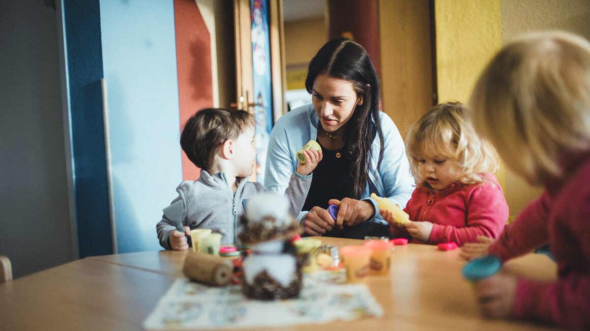 Eine Erzieherin arbeitet mit drei Kindern am Tisch, während sie mit Knete spielen.