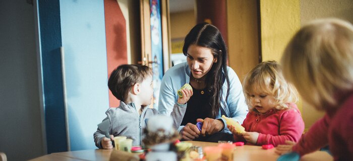 Eine Erzieherin arbeitet mit drei Kindern am Tisch, während sie mit Knete spielen.