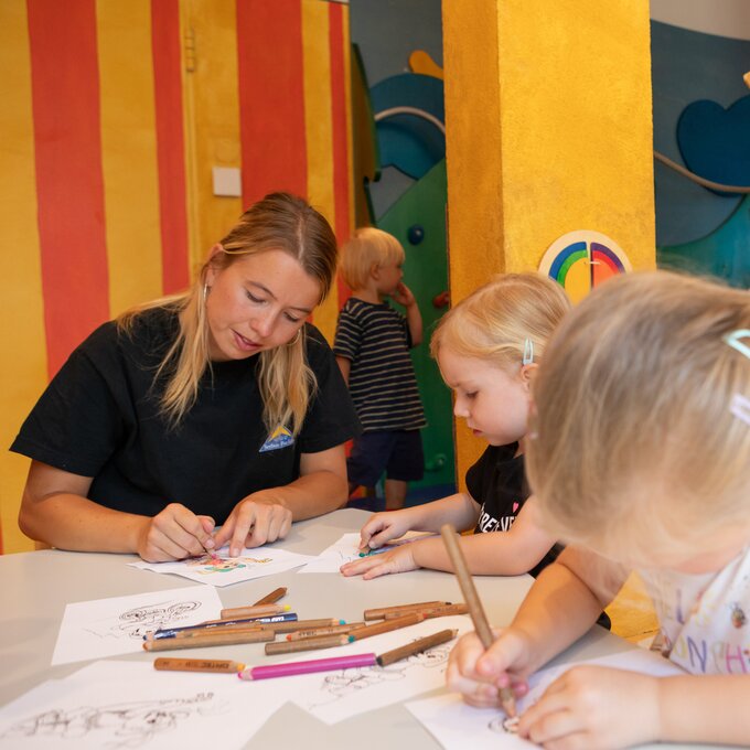 Een verzorger en twee kinderen zijn enthousiast aan het tekenen in een kleurrijke kamer.