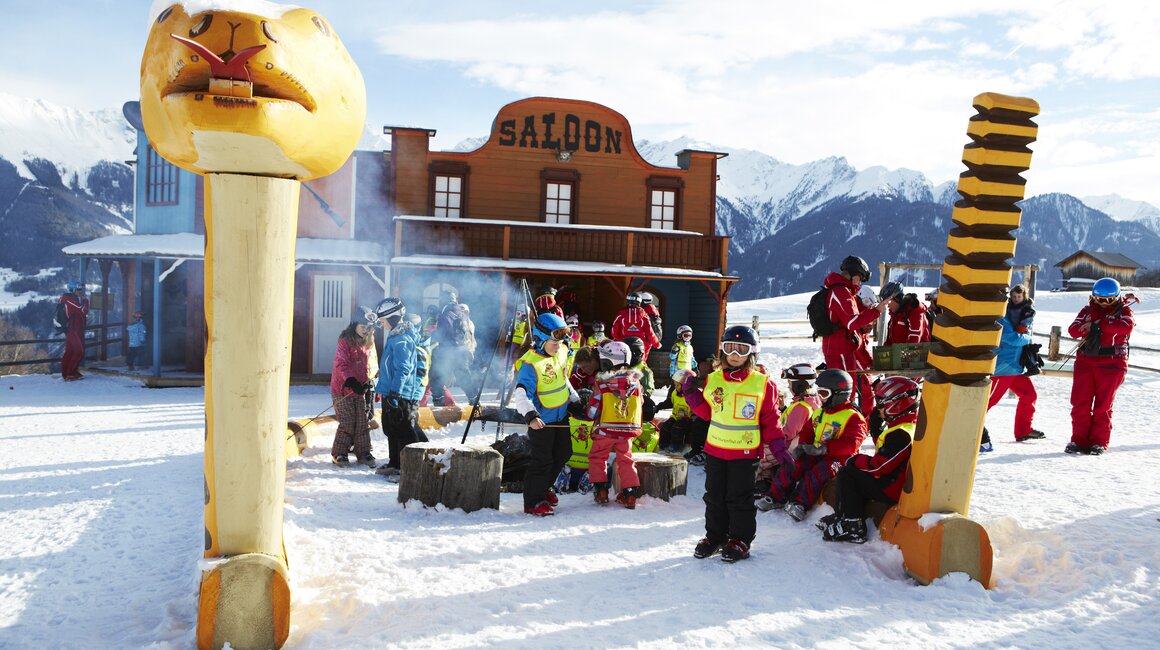Group of children in ski attire gathered in front of a saloon in the snow.