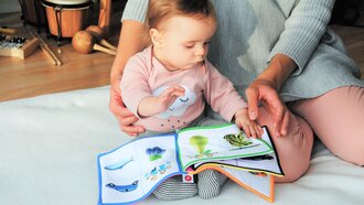 A baby sitting on the floor flipping through a colorful picture book.