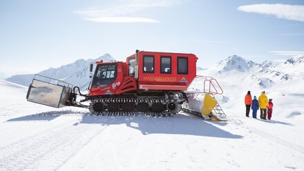 A red snow groomer stands in the snow overlooking the mountains.
