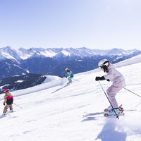 Drei Skifahrer auf einer schneebedeckten Piste mit Bergen im Hintergrund.