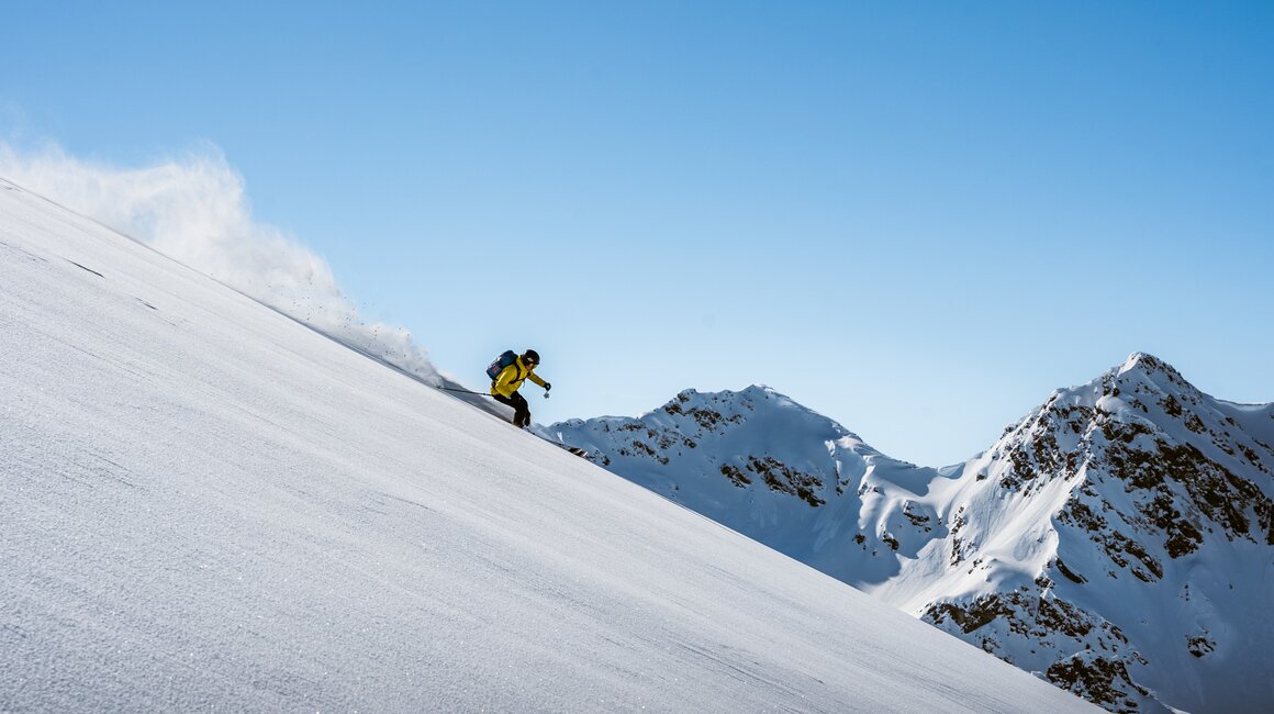Ein Skifahrer fährt die verschneite Piste hinunter unter blauem Himmel.