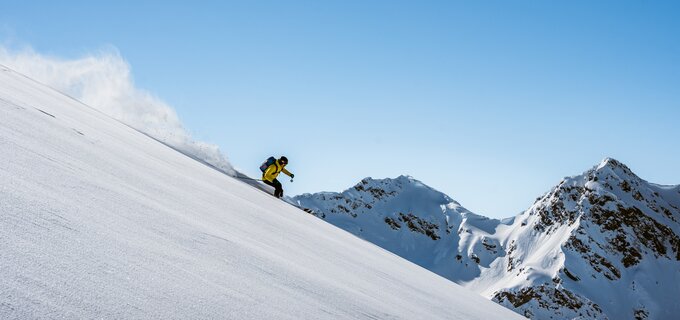 Een skiër daalt een besneeuwde helling af onder een heldere blauwe lucht.