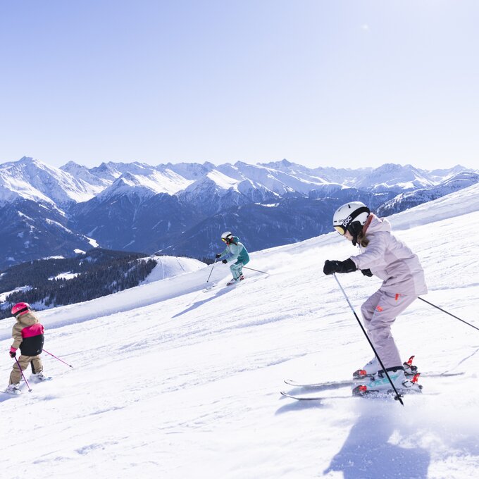 Drei Skifahrer auf einer schneebedeckten Piste mit Bergen im Hintergrund.
