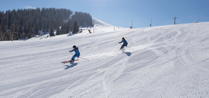 Zwei Skifahrer genießen eine sonnige Abfahrt auf einer verschneiten Piste.