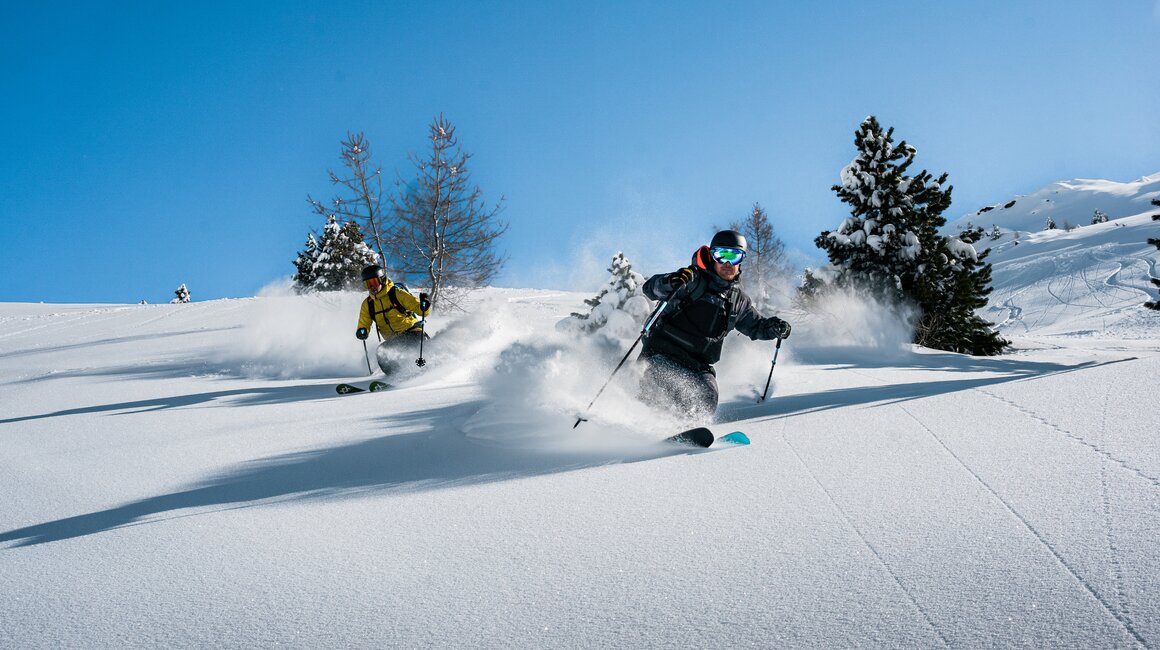 Zwei Skifahrer sausen durch frischen Pulverschnee auf einer Piste.
