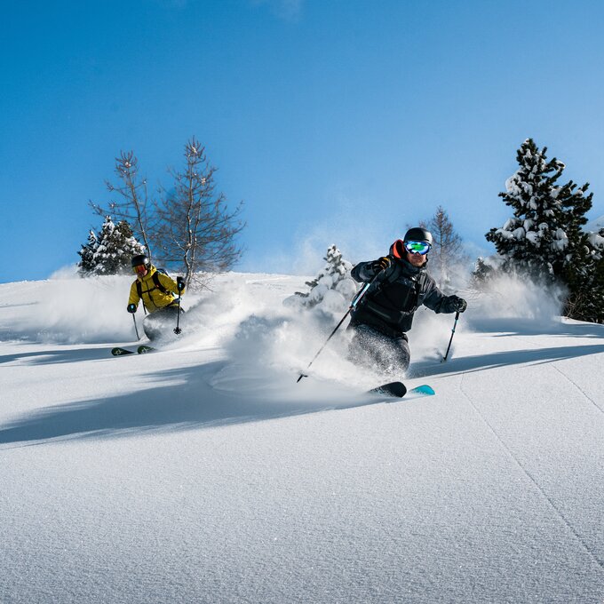 Twee skiërs glijden door verse poedersneeuw op een helling.