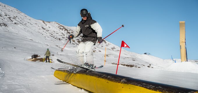 Skier with poles jumps over a yellow rail in the snow.