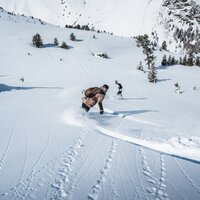 Zwei Personen snowboarden durch tiefen Pulverschnee in den Bergen.