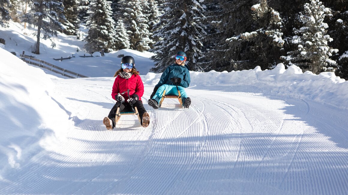 Zwei Kinder sitzen auf Schlitten in der verschneiten Landschaft.