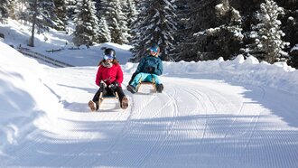 Zwei Kinder sitzen auf Schlitten in der verschneiten Landschaft.