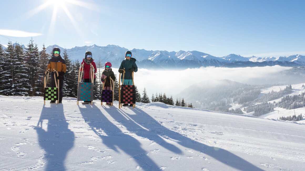 Familie steht auf dem Berg mit Skischlitten, umgeben von Schnee und Bergen.