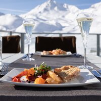 Ein Teller mit Speisen vor schneebedeckten Bergen und Gläsern Sekt.