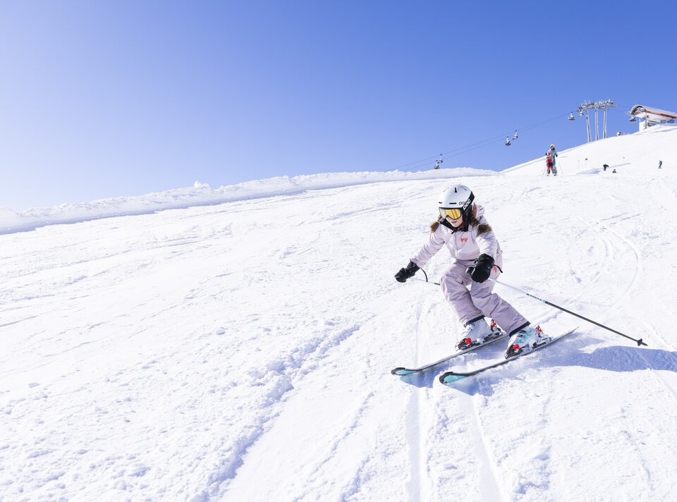 Ein Kind fährt Ski auf einer verschneiten Piste bei klarem Himmel.
