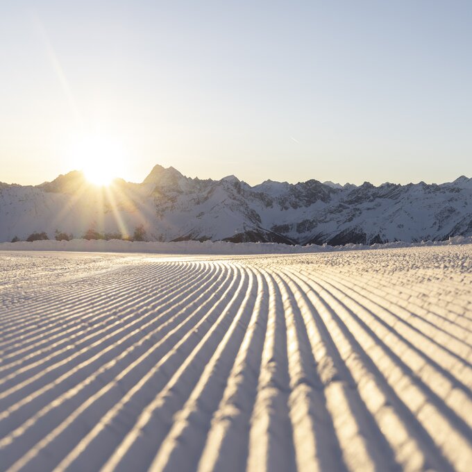 A sunrise casts light over snowy mountains with patterned snow trails.