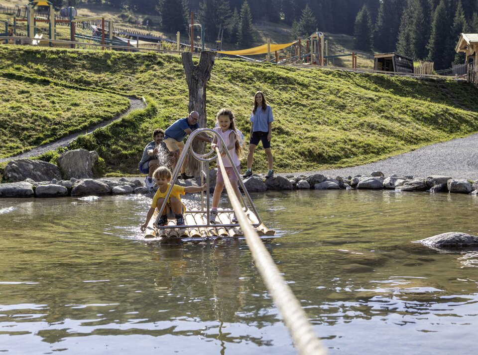 Zwei Kinder auf einem Floß im Wasser mit Eltern im Hintergrund.