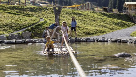 Zwei Kinder auf einem Floß im Wasser mit Eltern im Hintergrund.