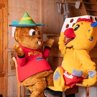 Zwei Maskottchen sitzen in einem Holzraum und unterhalten sich.
