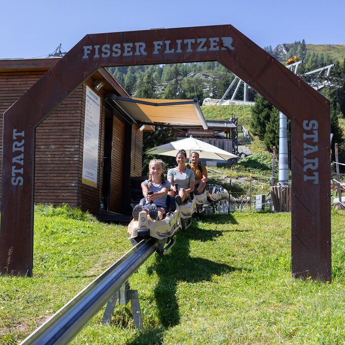 Zwei Personen fahren auf dem Fisser Flitzer, einer Sommerrodelbahn, bei sonnigem Wetter.