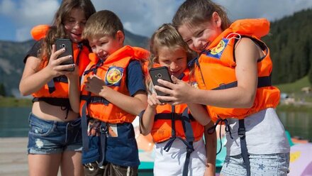Four kids in life jackets looking at their phones by the water.
