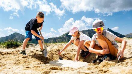 Three children digging in a sandy area with a shovel.