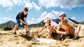 Drei Kinder graben in sandiger Umgebung mit Schaufel.