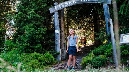 A smiling child stands under a wooden sign on a hiking path.
