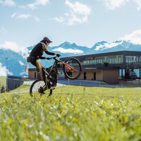 A mountain biker performs tricks on a lawn, with mountain scenery.