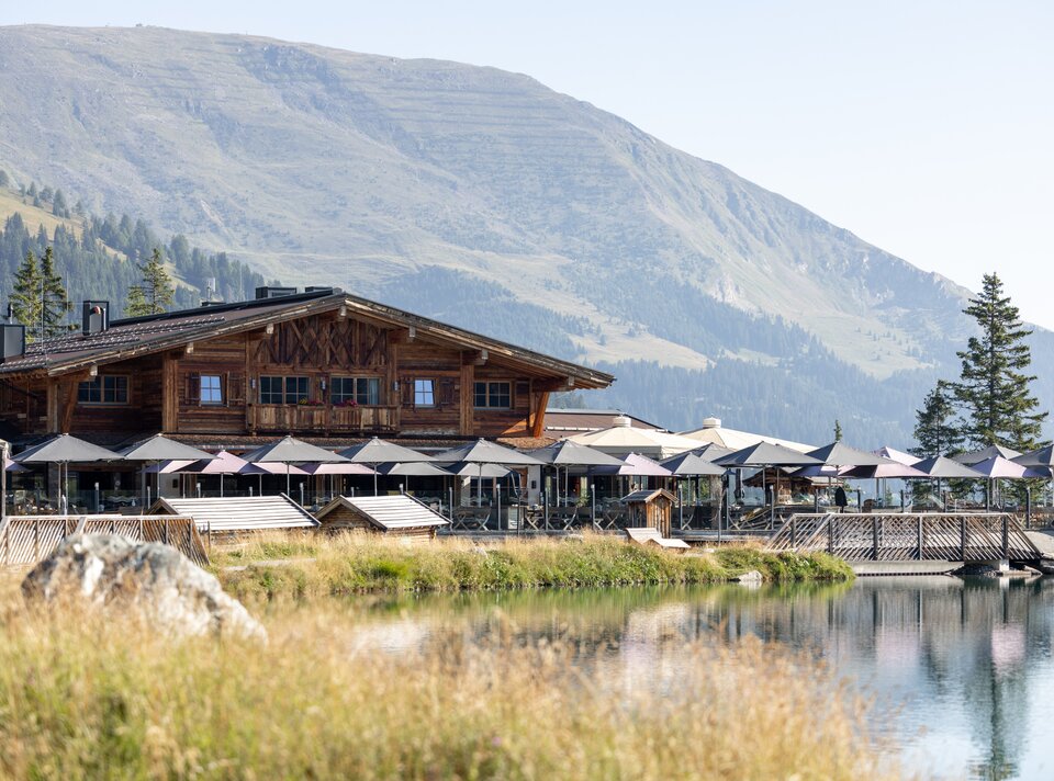 A wooden house with terraces by the lake and mountains in the background.