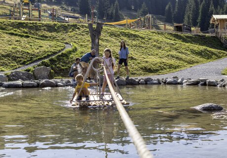 Zwei Kinder auf einem Floß im Wasser mit Eltern im Hintergrund.