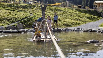 Zwei Kinder auf einem Floß im Wasser mit Eltern im Hintergrund.