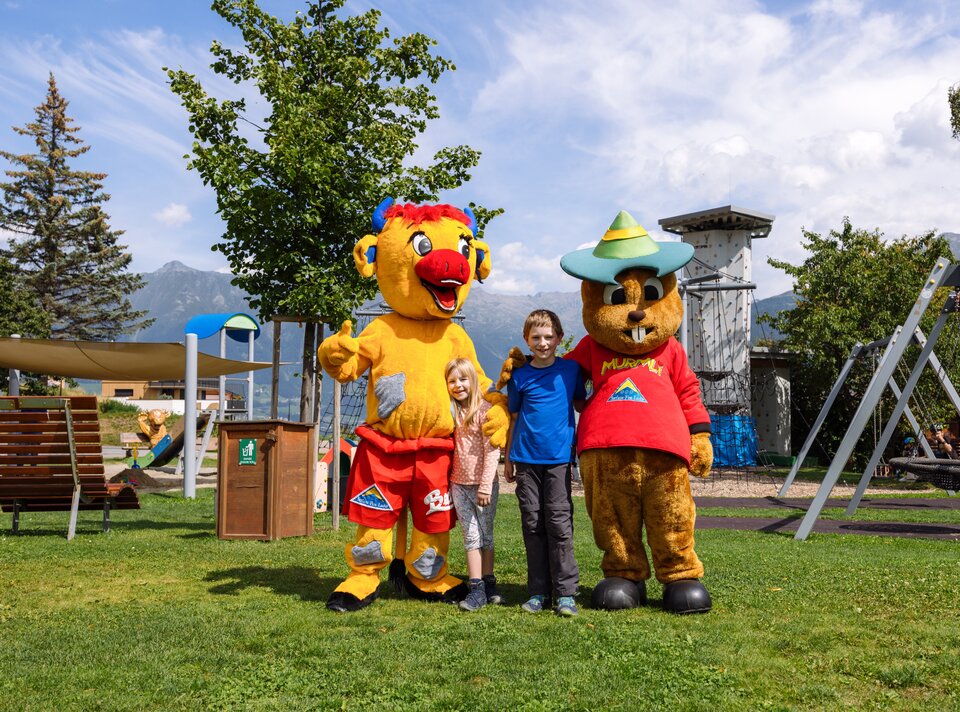 Twee kinderen poseren met twee dierenmaskottens in een park.