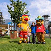 Zwei Kinder posieren mit zwei Tiermaskottchen in einem Park.