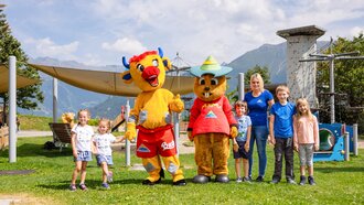 Groepsfoto met kinderen en maskotten in de buitenlucht, omringd door natuur.
