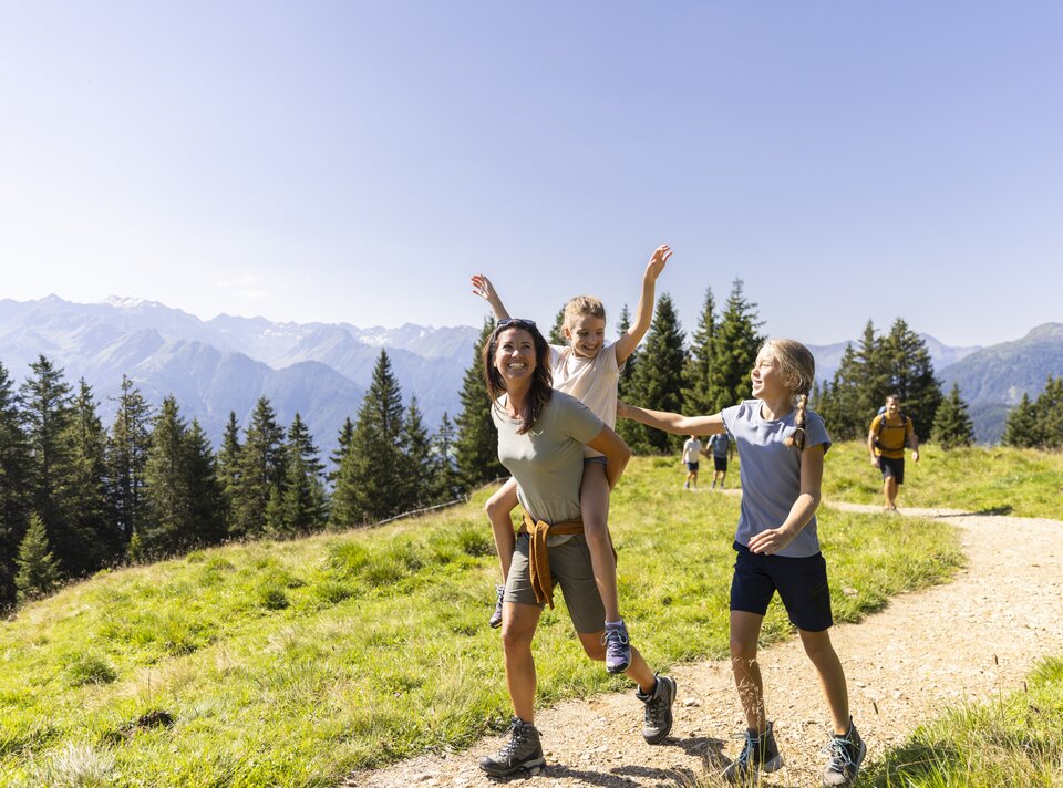 A woman carries a child on her back while another child walks beside her, surrounded by mountains.