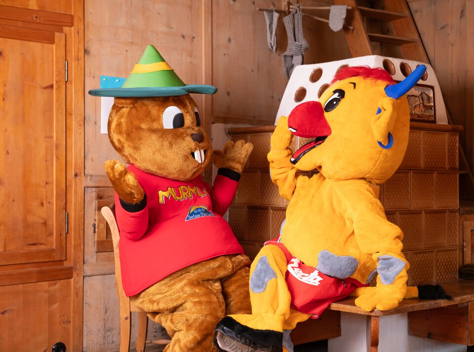 Zwei Maskottchen sitzen in einem Holzraum und unterhalten sich.