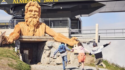 Three children happily run towards a large wooden sculpture.