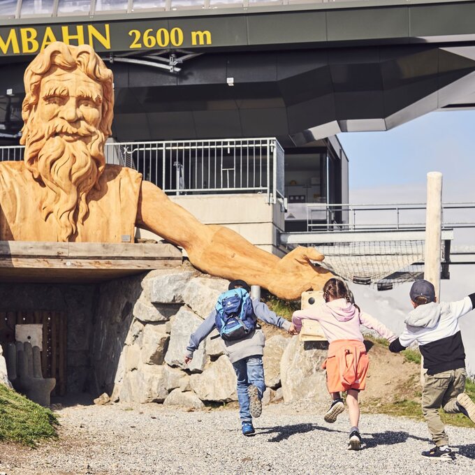 Three children happily run towards a large wooden sculpture.